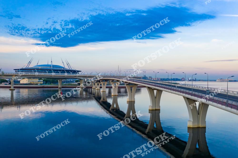 На фотографии изображён мост, ведущий к стадиону «Газпром Арена» на Крестовском острове. Архитектура моста гармонично сочетается с видом Финского залива и голубым утренним небом. Современный стадион стал символом спортивного развития Санкт-Петербурга. Отражения в воде придают пейзажу спокойствие и красоту.